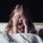 frightened kid screaming and covering eyes with hands while sitting on bedding near teddy bear